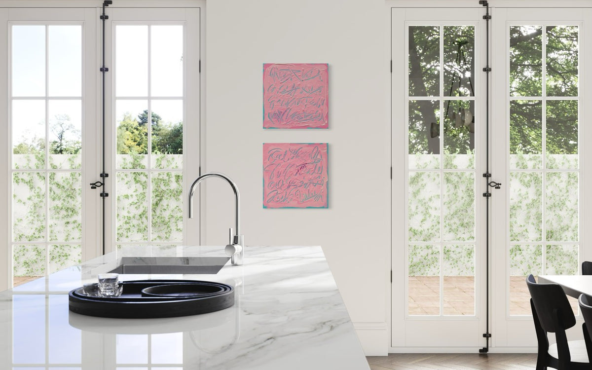 Two square pink abstract art prints with blue calligraphic patterns hang on a white wall in a bright kitchen with white French doors and a marble countertop.