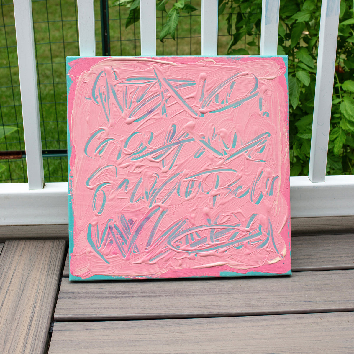 A square canvas painting with thick pink paint and abstract light blue and white brushstrokes, leaning against a white railing on a wooden deck with green foliage in the background.