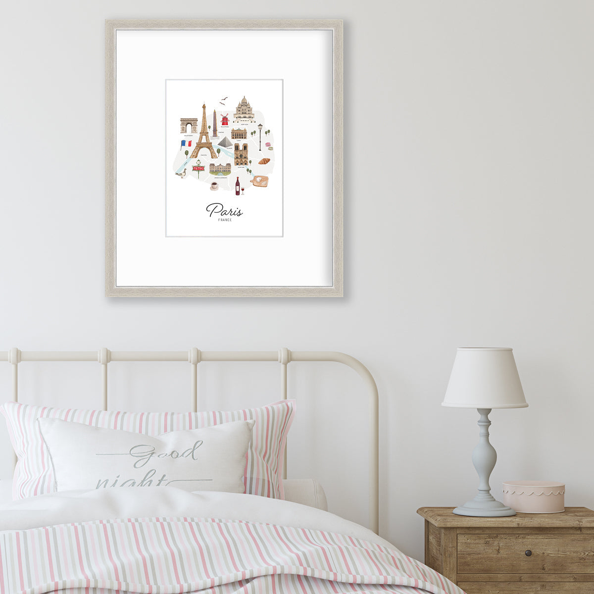 A multicolored map print of Paris, France with the famous landmarks in a silver frame with a mat hangs on a white wall above a white iron headboard, pink and grey bedding and a wood night stand with a small lamp and a light pink decorative box.