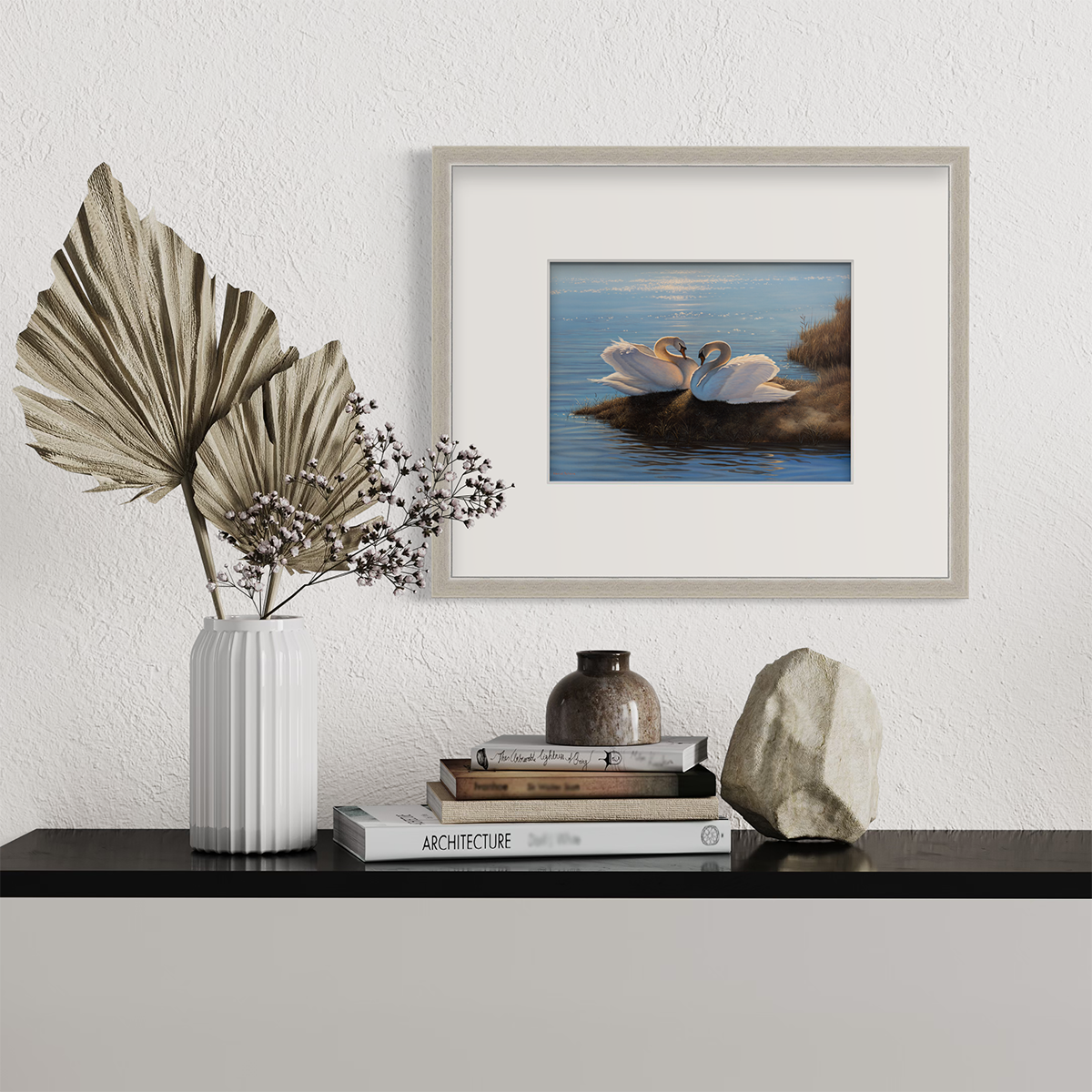 A print of two swans sitting by a body of water. Hangs in a silver frame above a black countertop with a stack of books and a vase with decorative stems..