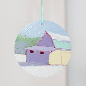 A hand-painted ornament depicting a barn in a winter landscape hangs from a light blue ribbon in front of a white wall. 