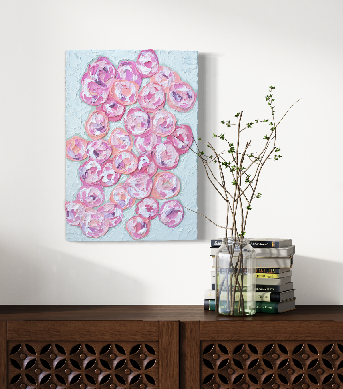 A very textured abstract painting with celadon background and circle type abstract flowers in pink, peach, plum and white hangs on a white wall above a credenza with books and a glass vase holding branches with green buds.