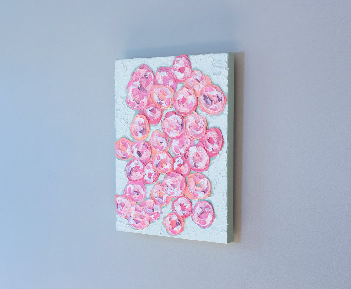 A very textured abstract painting with celadon background and circle type abstract flowers in pink, peach, plum and white hangs on a grey wall.