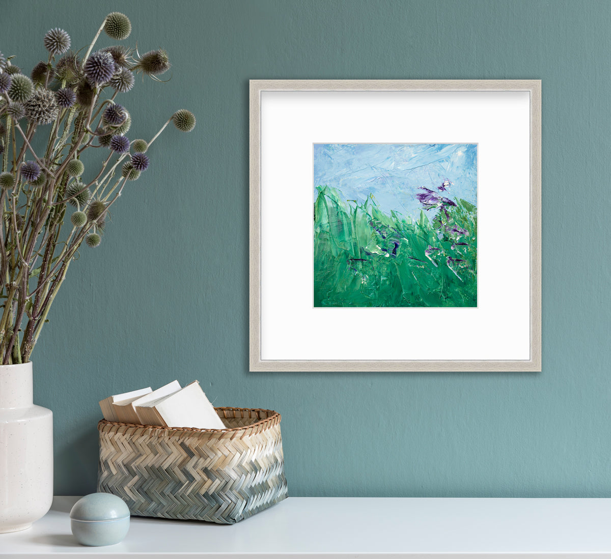 A blue, green and purple abstract landscape print in a silver frame with a mat hangs on a teal wall above a white surface with a beige and teal woven basket with card stock, a small round teal trinket and a white vase with green and purple pom pom stems.