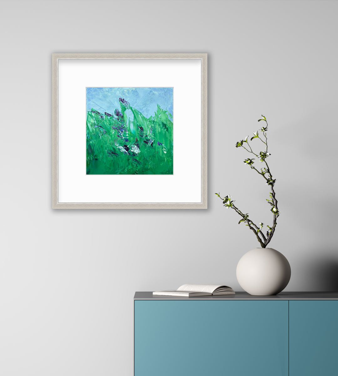 A blue and green abstract landscape print in a silver frame with a mat hangs on a white wall above a teal cabinet with an open book and a white rounded vase with greenery stems.