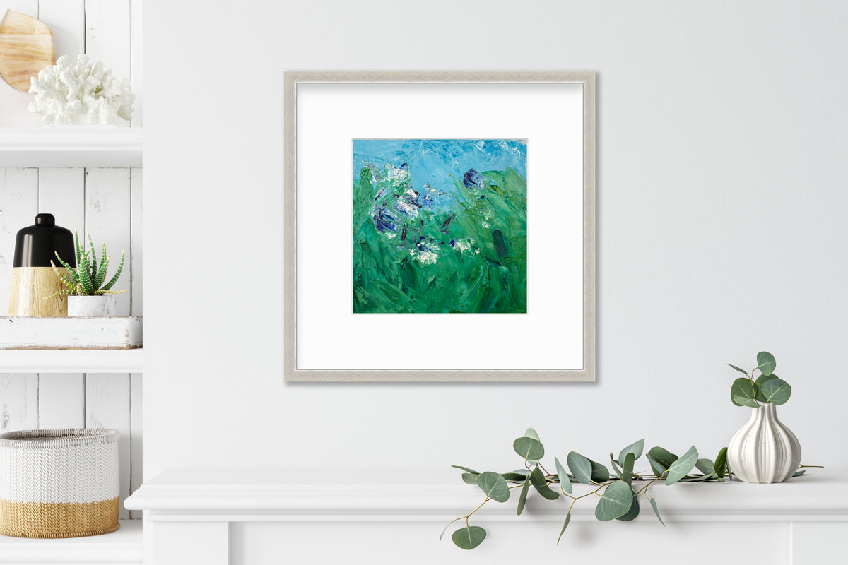 A blue, green, purple and white abstract landscape print in a silver frame with a mat hangs on a white wall above a white shelf with a eucalyptus filled white vase and next to a built in bookshelf with accessories.
