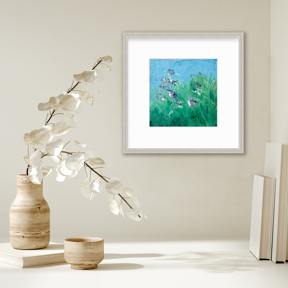 A blue, green, purple and white abstract landscape print in a silver frame with a mat hangs on a white wall above a stack of books and a wood vase with white leaf stems.