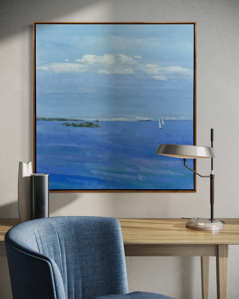 Framed painting of a blue landscape with a sailboat on a wall above a desk with a lamp and chair.