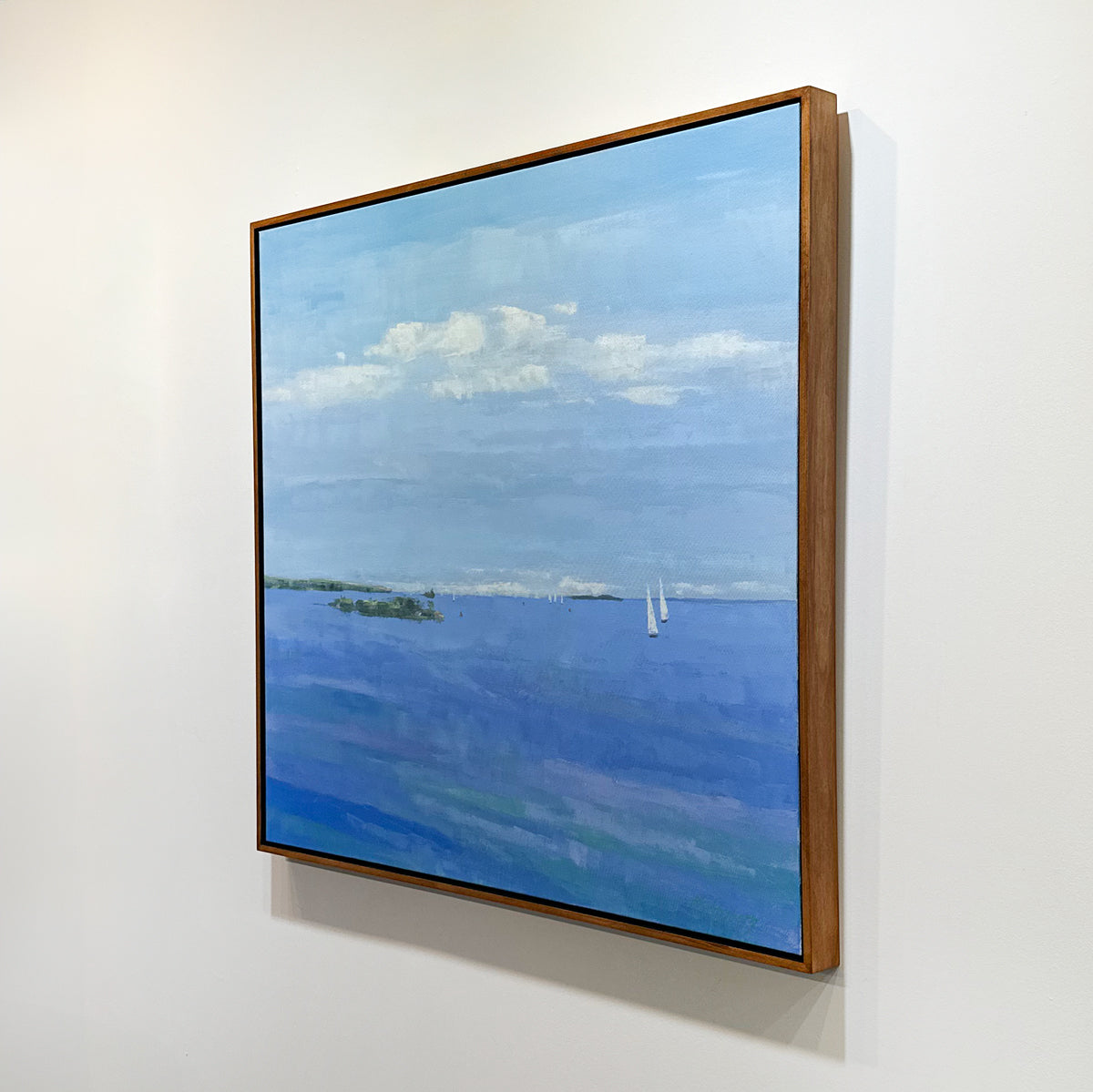 Side view of a framed painting of a blue landscape with clouds on a white wall.