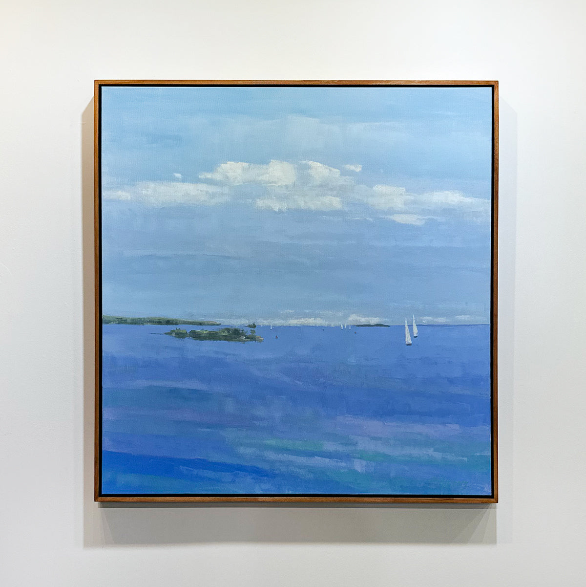 Framed painting of a blue landscape with clouds and sailboats on a white wall.
