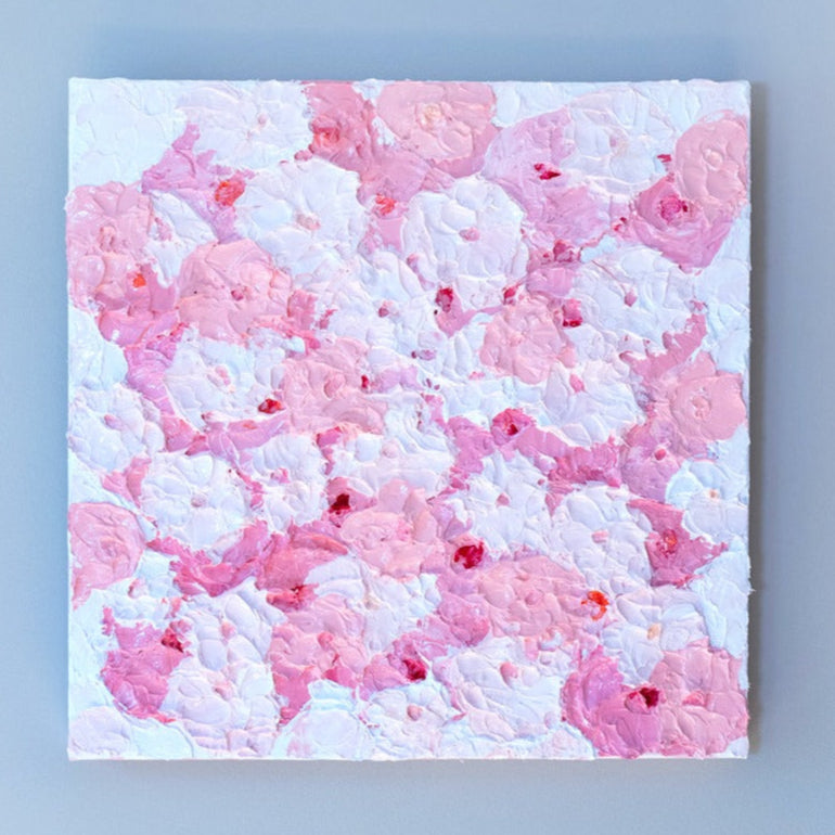 A very textured abstract painting with white background and circle type abstract flowers in pink and white hangs on a grey wall.