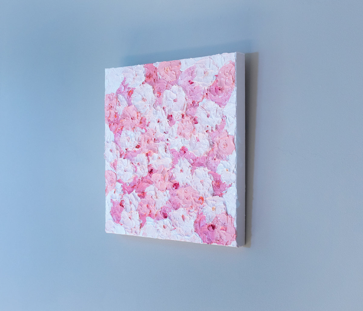 A very textured abstract painting with white background and circle type abstract flowers in pink and white hangs on a grey wall, side view.