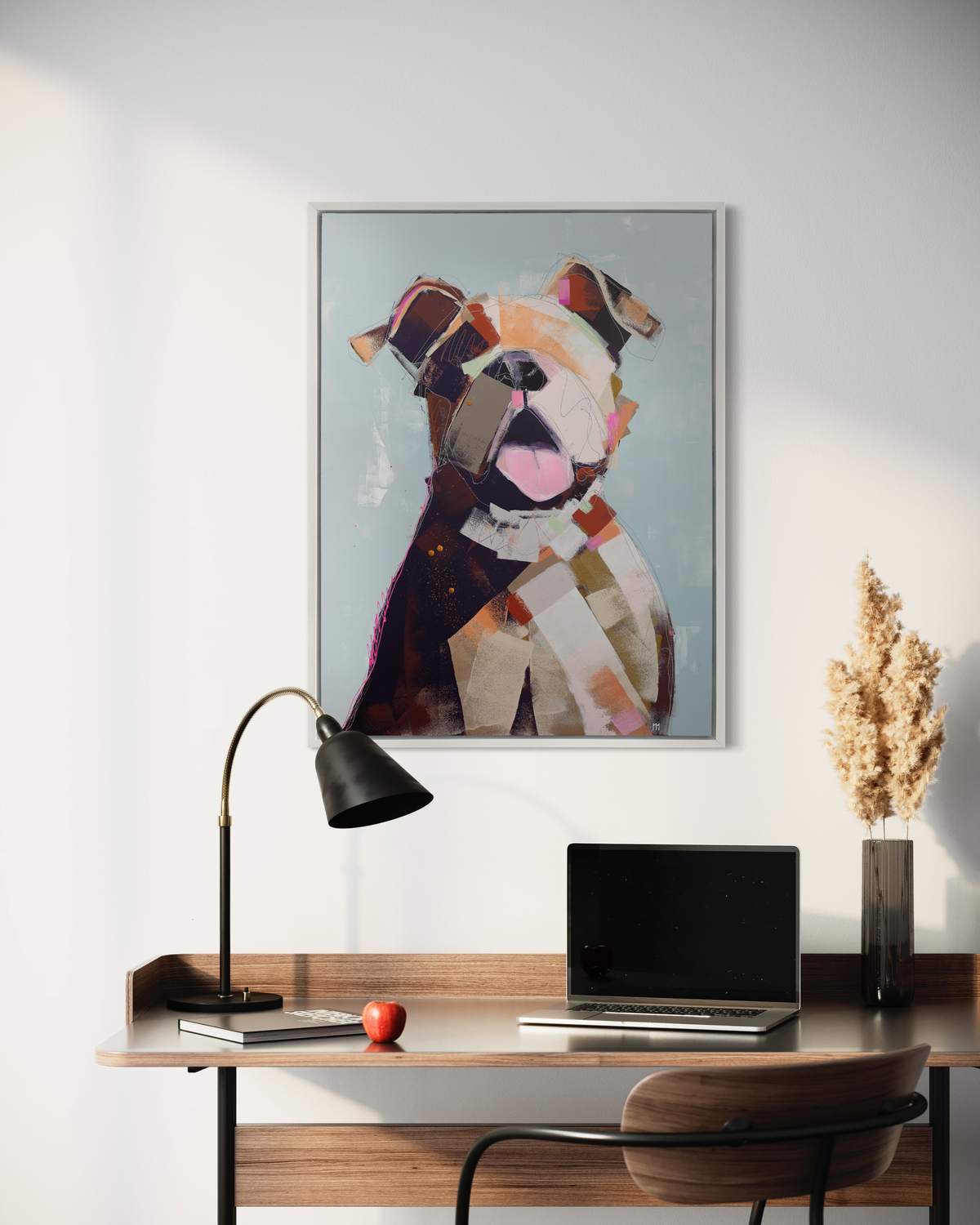 A light blue, orange, brown, pink and navy abstracted dog print in a white floater frame hangs on a white wall above a wood desk with matching chair, a laptop, a thin vase with wheat stems and a black desk lamp.