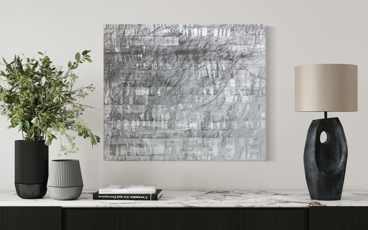 A black and silver abstract painting hangs on a white wall above a marble console table with black and white table decor and a modern lamp. 