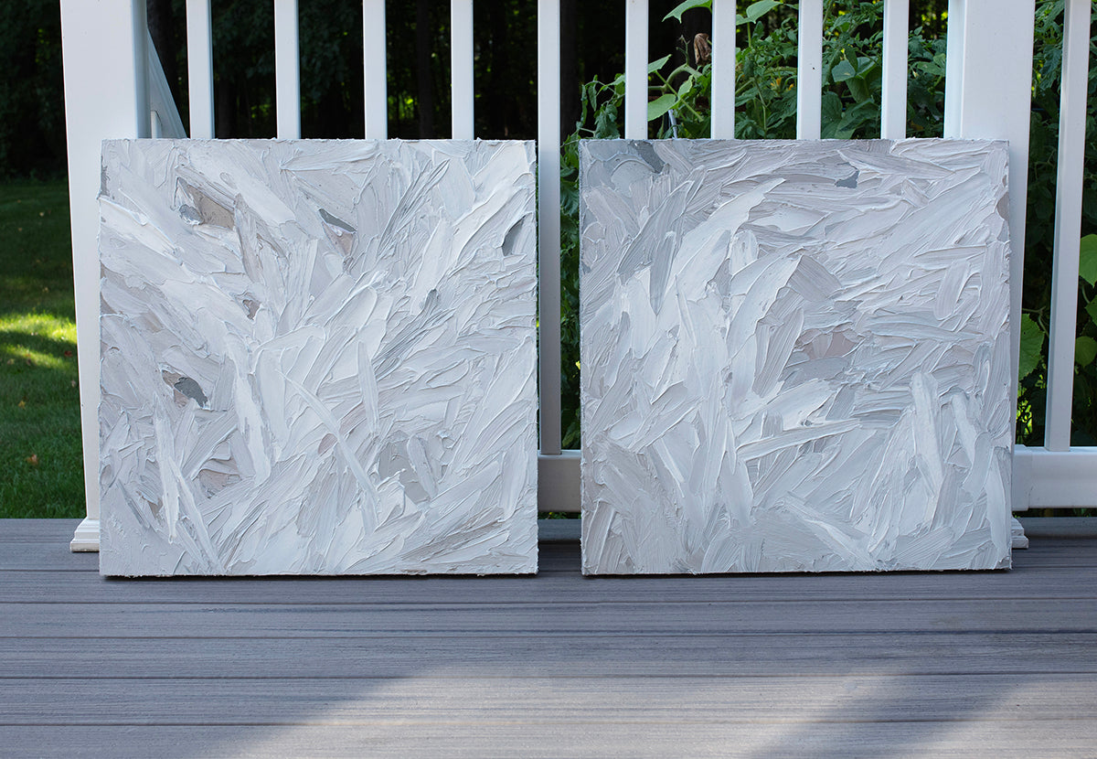 A pair of thickly textured paintings in grey, white, blue grey and plum grey by Teodora Guererra leaning outside on a deck.