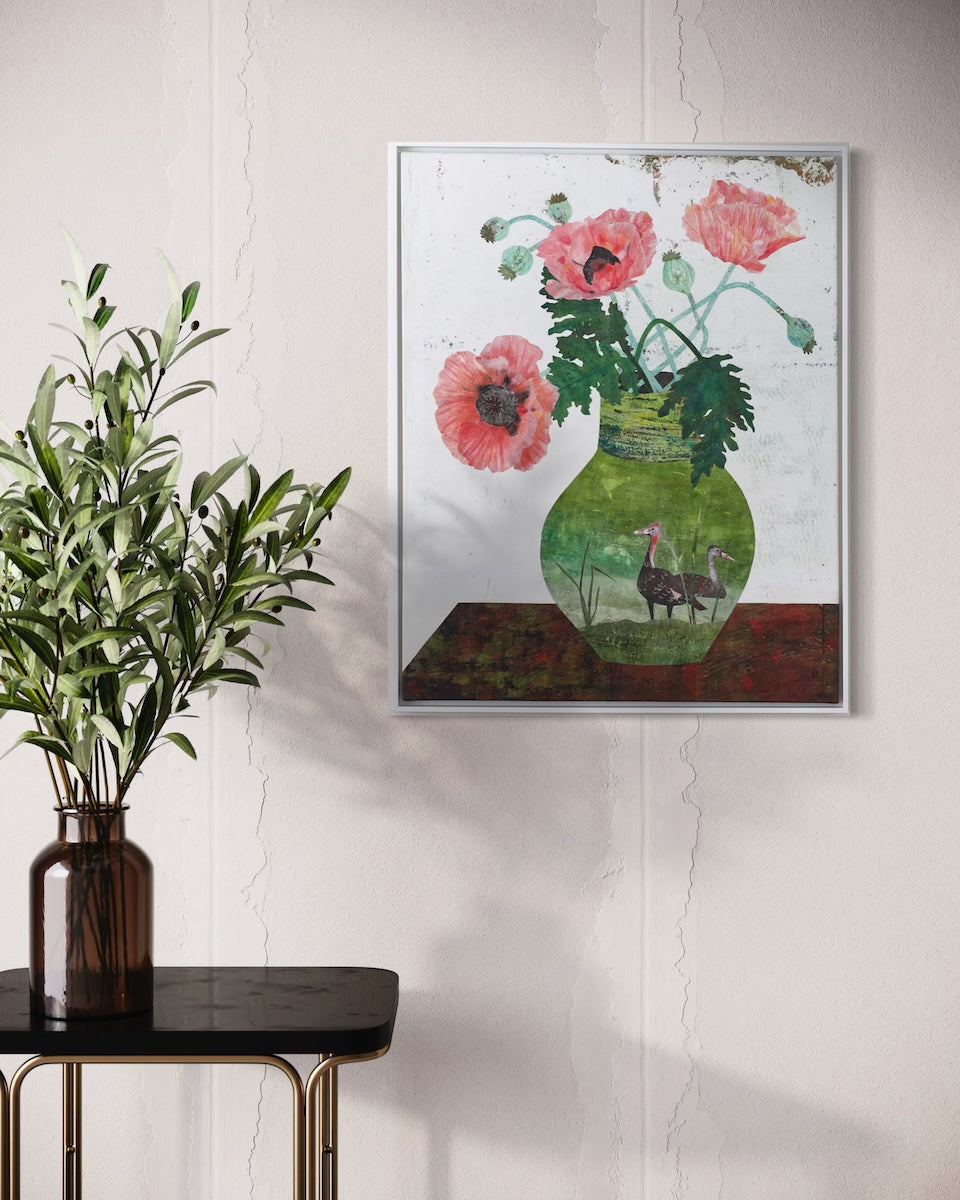 A framed collage painting depicting pink poppies in a green vase hanging on a textured off-white wall, styled next to a dark brown glass vase with green leaves on a black side table.