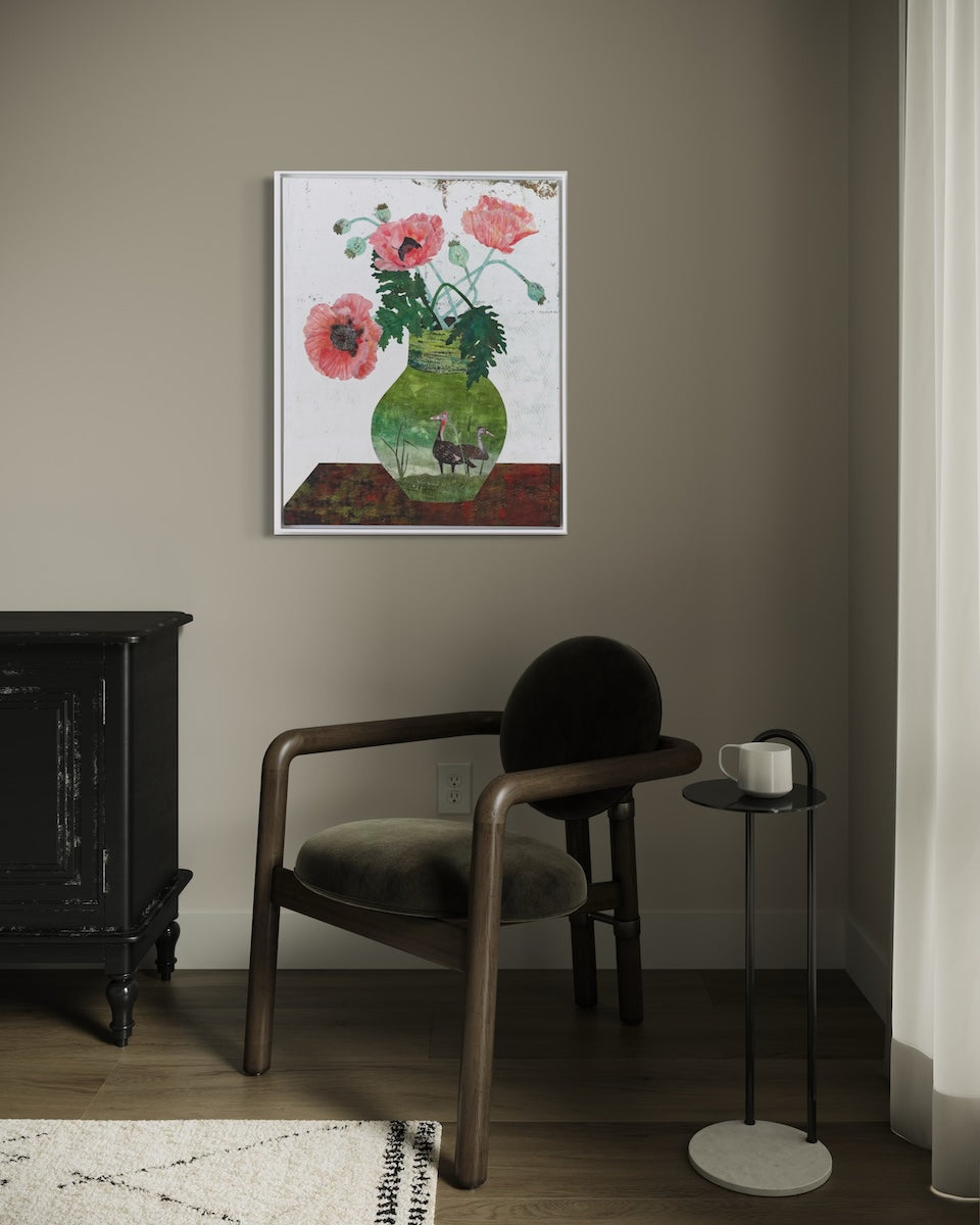 A framed collage painting depicting a green glass vase filled with pink poppies, hanging on a taupe-gray wall in a modern living room setting with a wooden armchair and a side table.