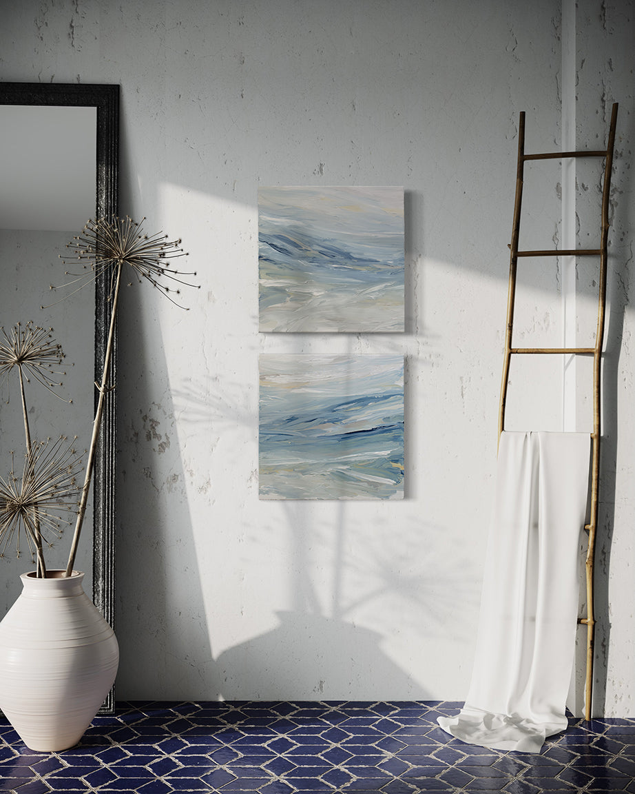 A pair of abstract coastal paintings with blue, grey, white, cream, stone, teal, and yellow ochre paint hanging on a white  stucco wall by Teodora Guererra. To the right is a natural wood decorative ladder with a white fabric draped over it. to the left is a wall mirror with a dark ridged frame leaning on the wall and a large white ceramic pot with decorative sticks in front. The floor below is a dark blue tile in a geometric pattern with grey grout.