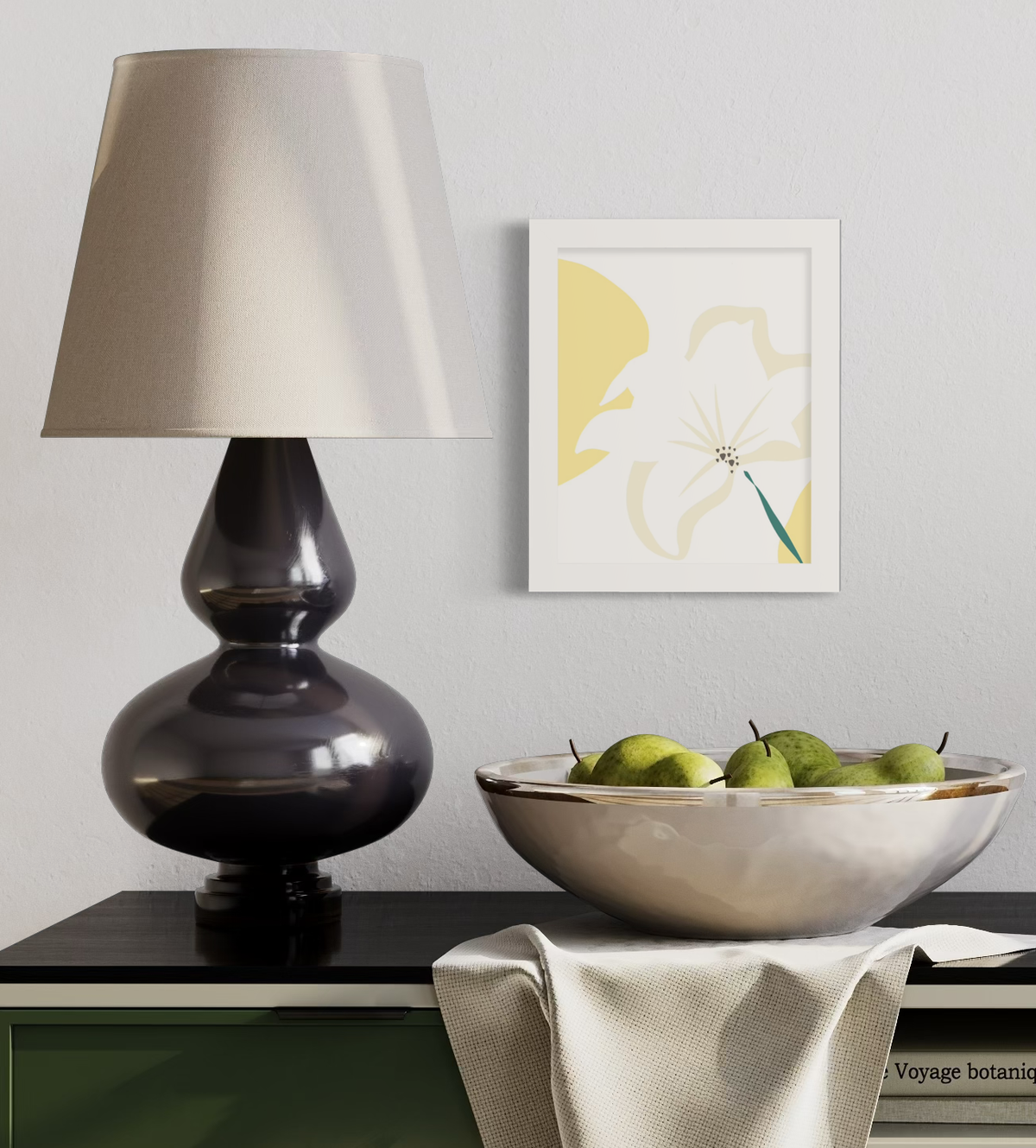 A light yellow abstract botanical art print in an unmatted white frame hangs on a white wall above a black surface with a bowl of pears and a dark modern lamp.