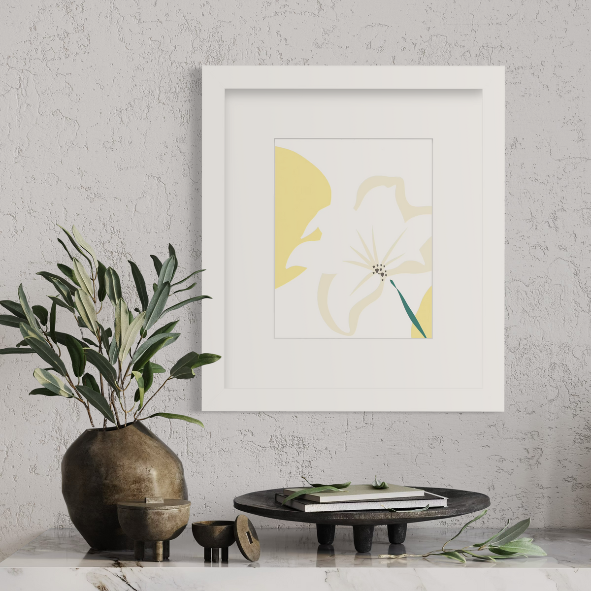 A light yellow abstract botanical art print in a white frame with a mat hangs on a light grey wall above a marble surface with a dark wood tray, small decorative containers and a dark misshapen vase with greenery.
