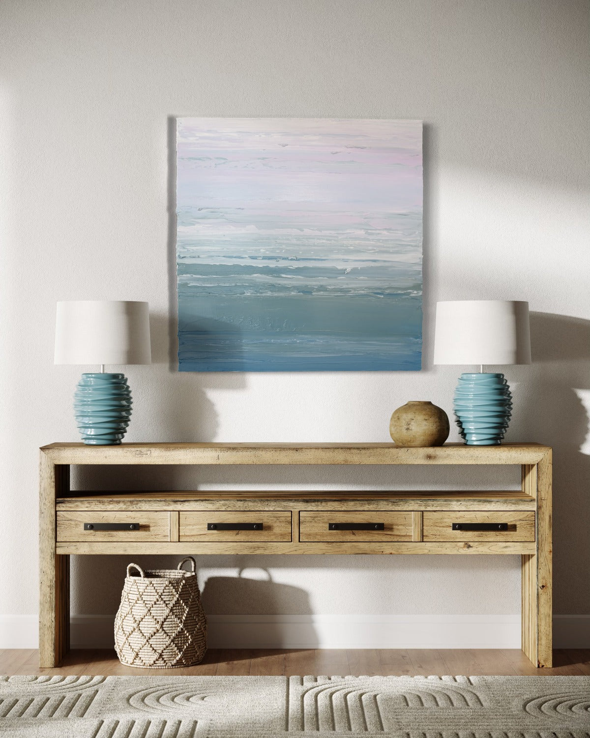 A teal blue, teal green, lavender and pale lavender textured abstract painting by Teodora Guererra hangs on a wall above a driftwood console with a basket below and 2 teal ceramic lamps on either side.