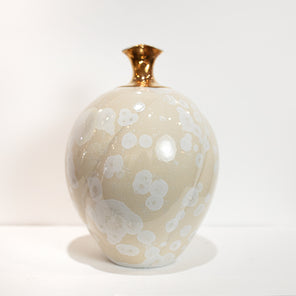 A cream colored vessel with white crystalline glaze and a narrow gold neck rests on a white surface in front of a white wall. 