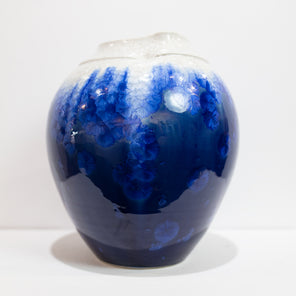 A blue and white glazed ceramic vase rests on a white surface in front of a white wall. 