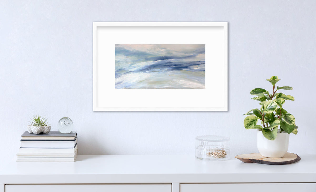 A blue, white and light green abstract print in a white frame with a mat hangs on a white wall above a white surface with a stack of books, a round clear container with gold accessories and a potted plant.