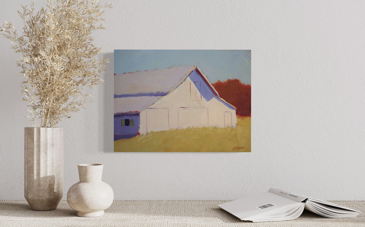 A small landscape painting of a barn hangs on a white wall above a table surface with stone vase decor and an open book facing down. 