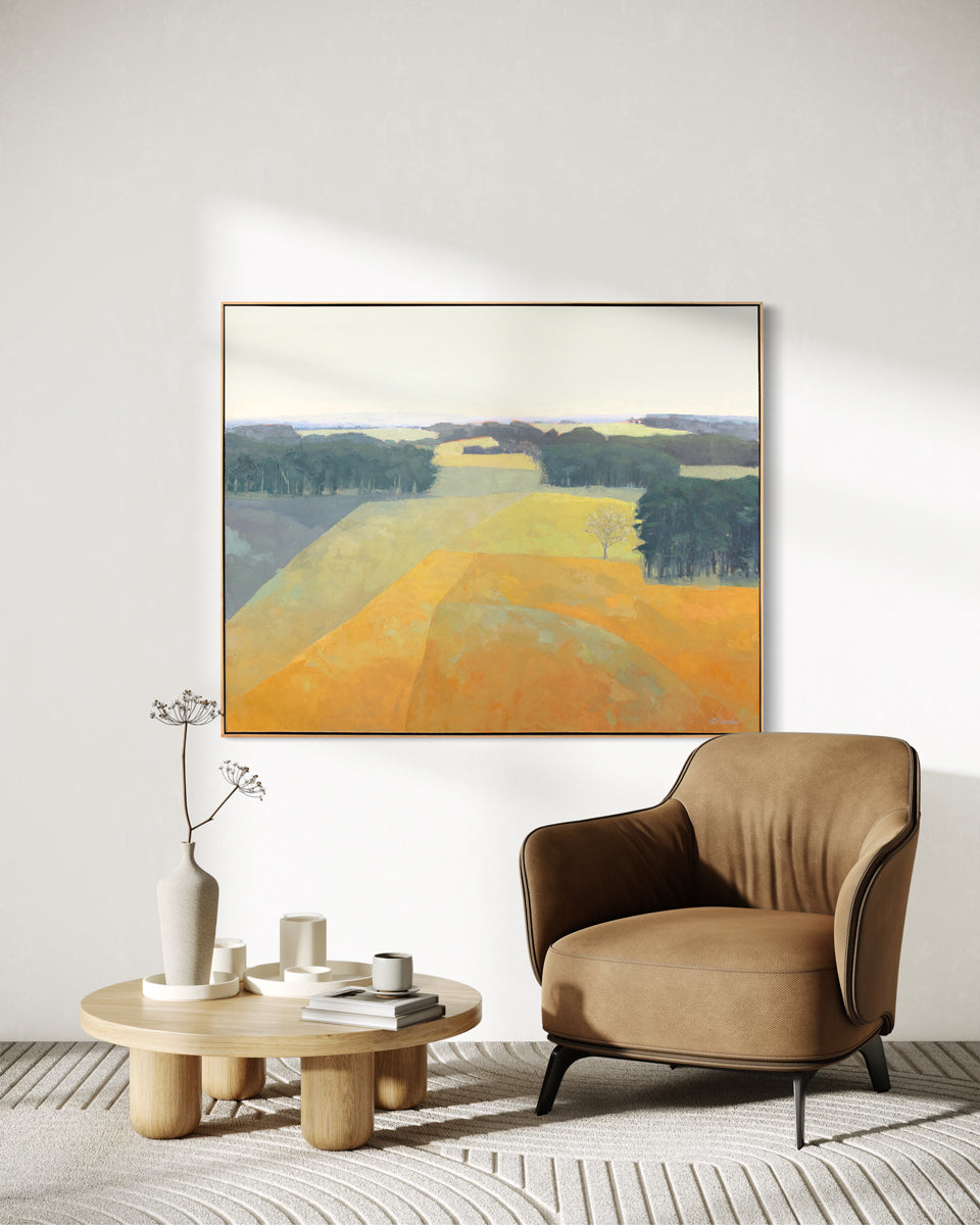 A yellow and green abstracted landscape painting hangs on a white wall above a brown suede armchair and a low wooden coffee table with minimalistic serveware and decor. 