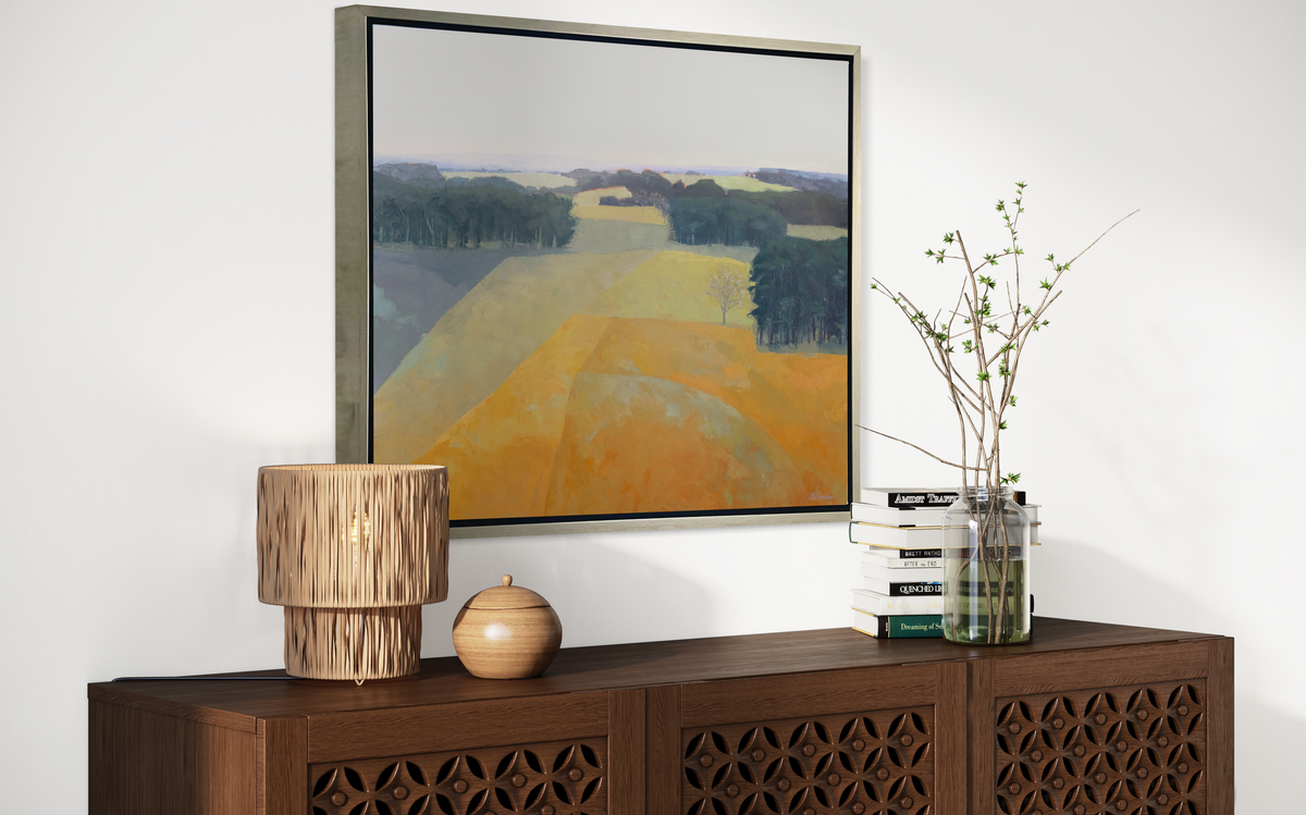 A side view of a light blue, green and light orange abstract landscape print in a silver floater frame hangs on a white wall above a dark wood credenza with a stack of books, a clear jar with greenery stems and decorative accessories.