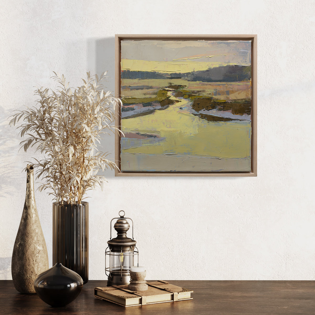 A framed yellow and brown abstracted landscape painting hangs on a white wall above a wood table surface with rustic table decor. 
