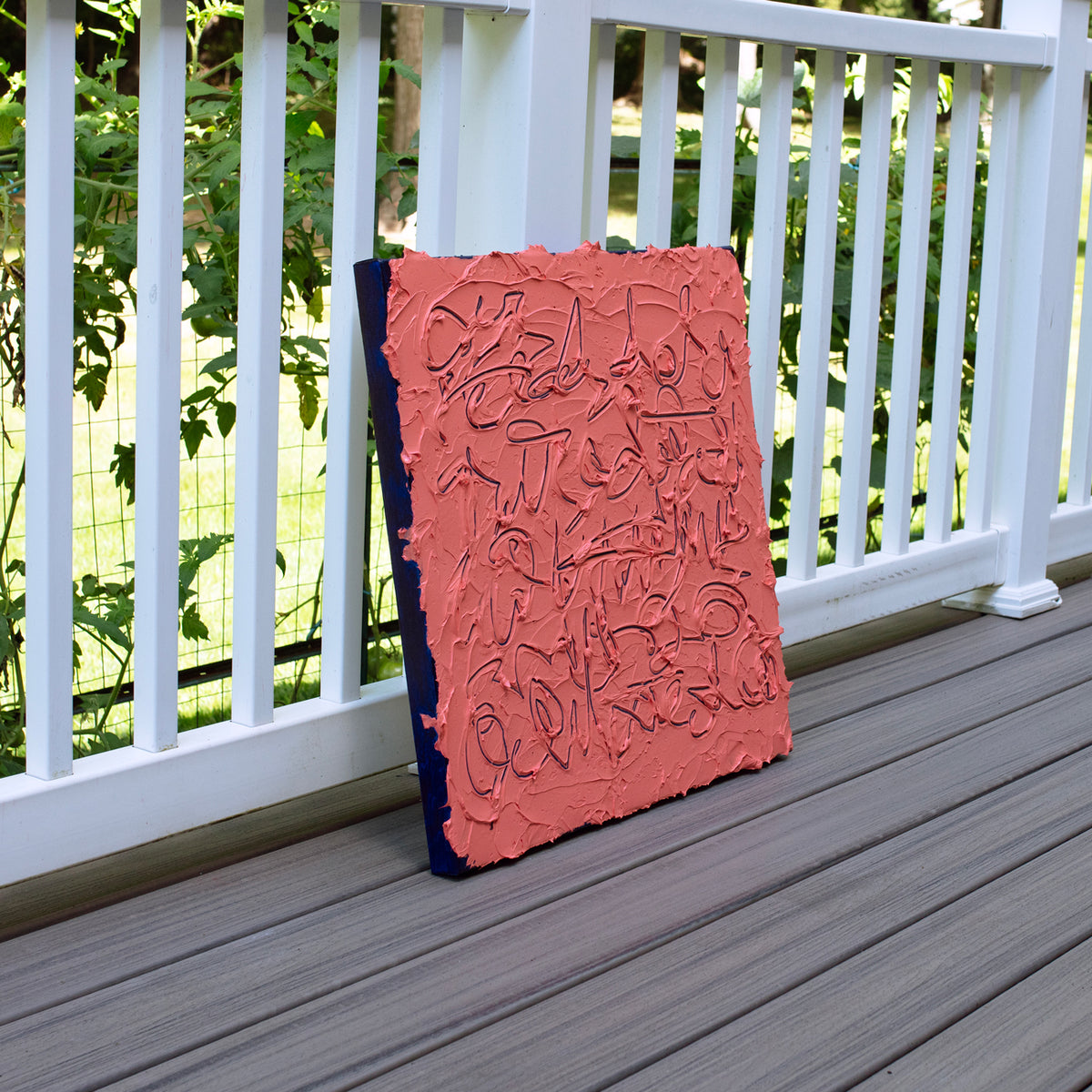 An abstract textured canvas painting in terracotta pink and dark navy blue, leaning against a white wooden railing on a gray composite deck.