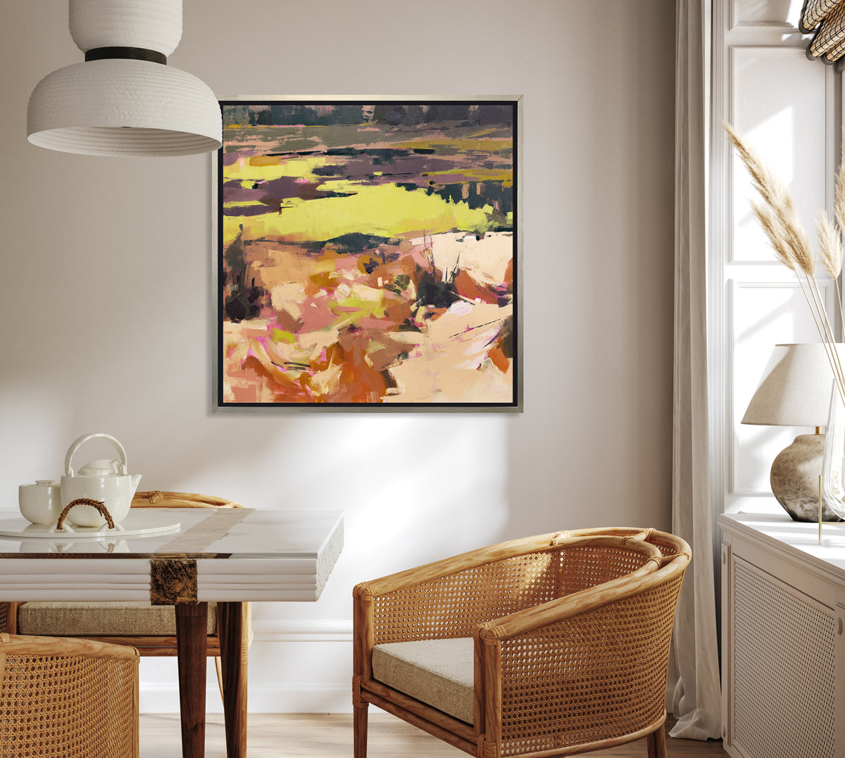 A yellow and orange abstracted landscape art print framed in a silver frame hangs on a light wall above a white dining table with wicker chairs and white decor. 