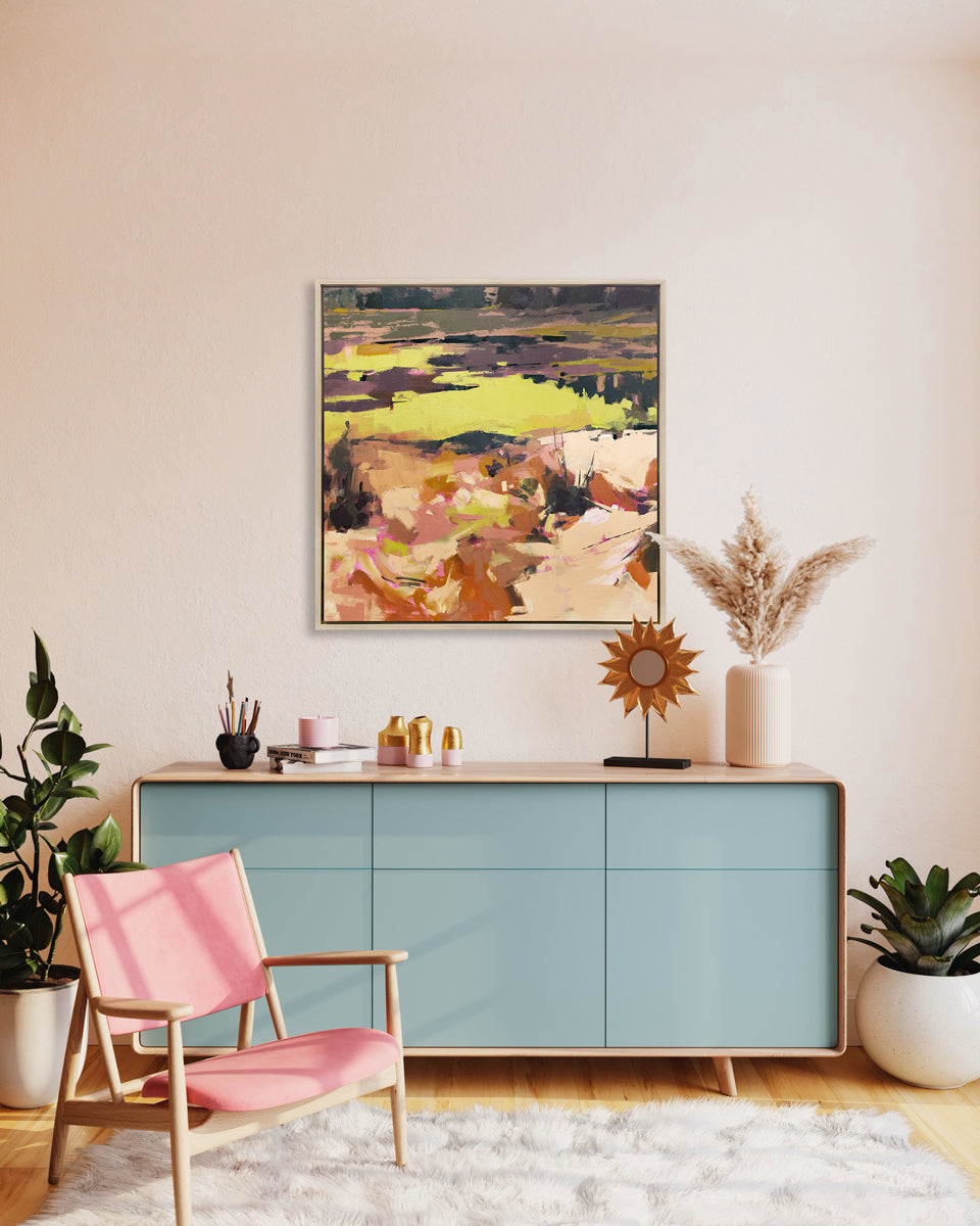 A yellow and orange abstracted landscape art print framed in a silver frame hangs on a white wall above a light blue console table and a pink armchair with eclectic table decor and potted plants. 