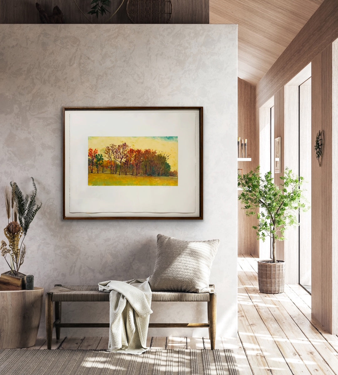 Framed painting of a landscape with trees and a yellow sky on a white wall hanging over a bench with a pillow and throw.