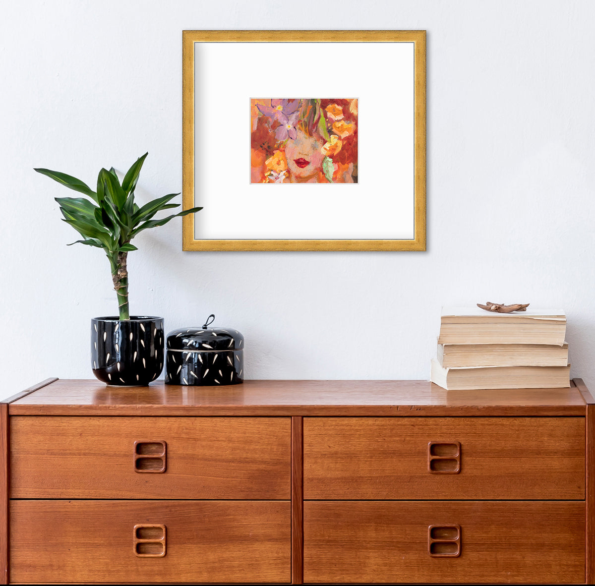 A red, orange, yellow and purple figural print in a gold frame with a mat hangs on a white wall above a wood cabinet with a stack of books, a potted plant in a black and white planter and a small matching black and white canister with a lid.