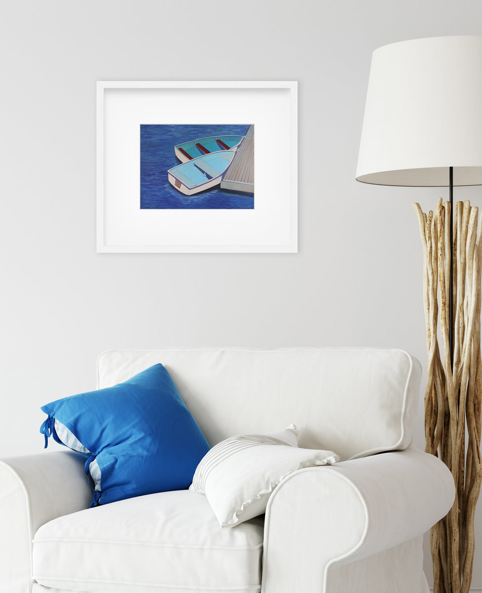 A blue, light blue and beige contemporary nautical print of two docked rowboats in a white frame with a mat hangs on a white wall above a white arm chair with a blue pillow and a lamp to the right. 