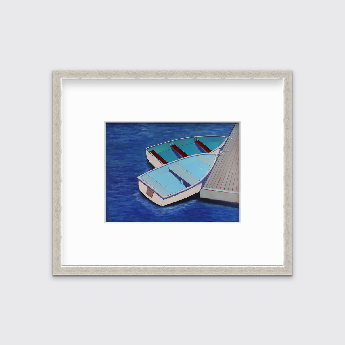 A blue, light blue and beige contemporary nautical print of two docked rowboats in a silver frame with a mat hangs on a white wall.