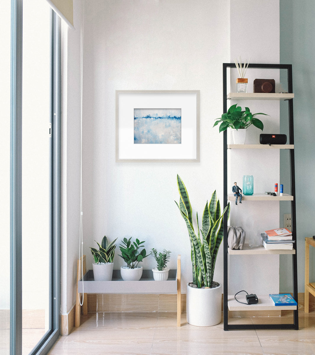 An abstract blue and white print hangs in an entry way. The print is next to a tall shelf and over multiple plants.