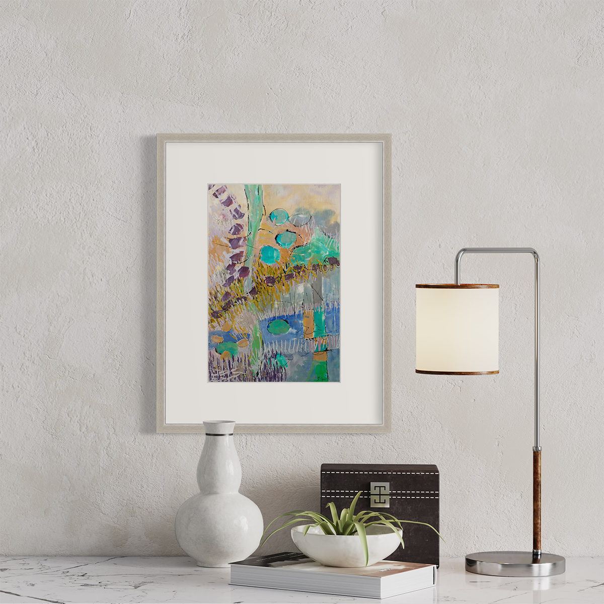 A teal, orange, purple and yellow abstract landscape print in a silver frame with a mat hangs on a white wall above a white marble surface with a small lamp, a black box, a book and decorative vases.