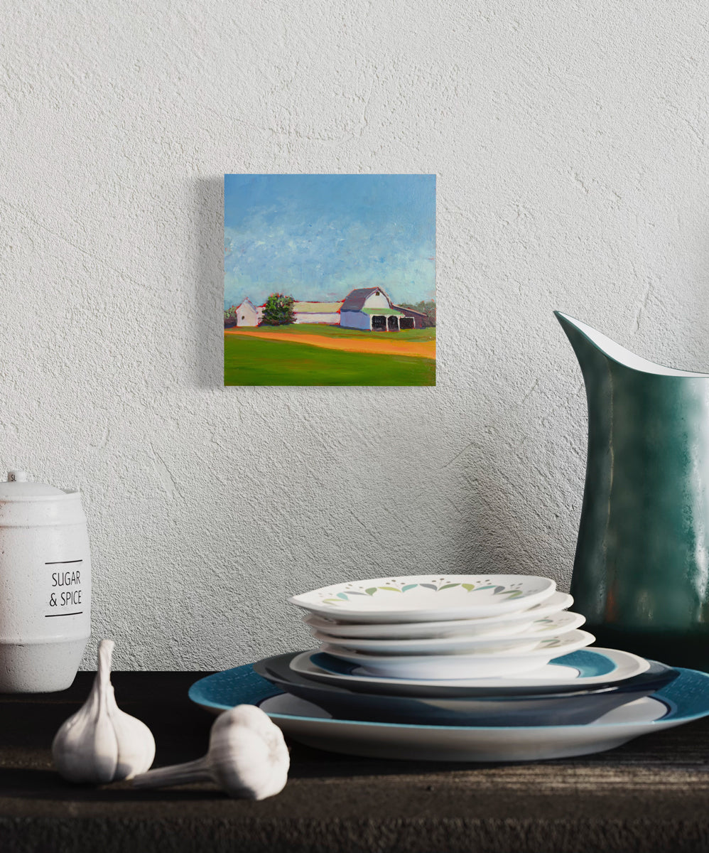 A blue, green, and orange landscape painting hangs on a textured wall above a stack of white and blue plates, a sugar bowl, cloves of garlic and a teal water pitcher. 