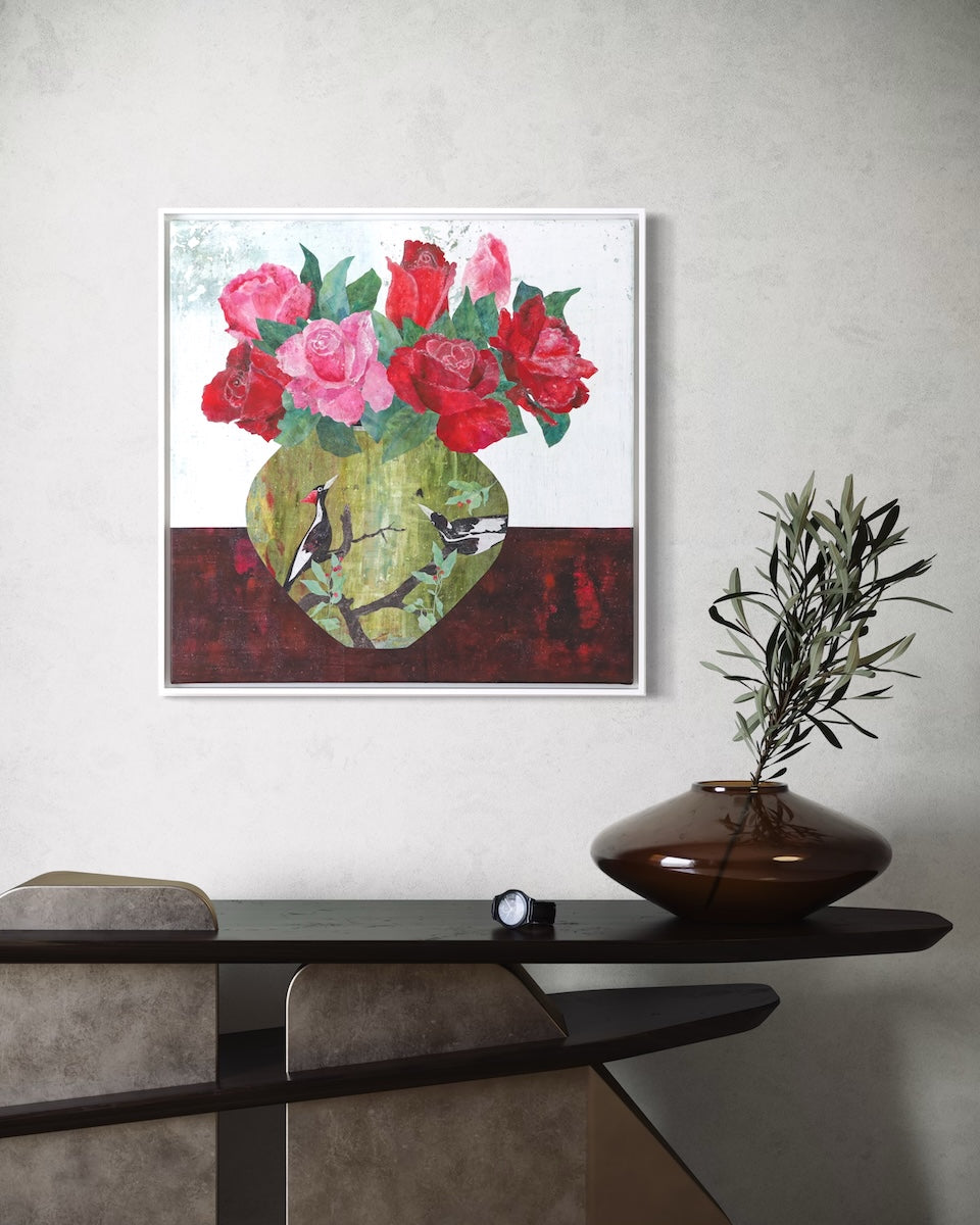 A framed collage painting of red and pink roses in a green vase with birds, displayed on a textured light gray wall above a dark console table with a brown vase and greenery.