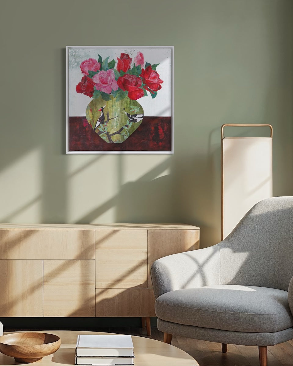 A framed floral collage hanging on a sage green wall above a light wood sideboard, with a gray armchair and a floor lamp in a sunlit living room setting.