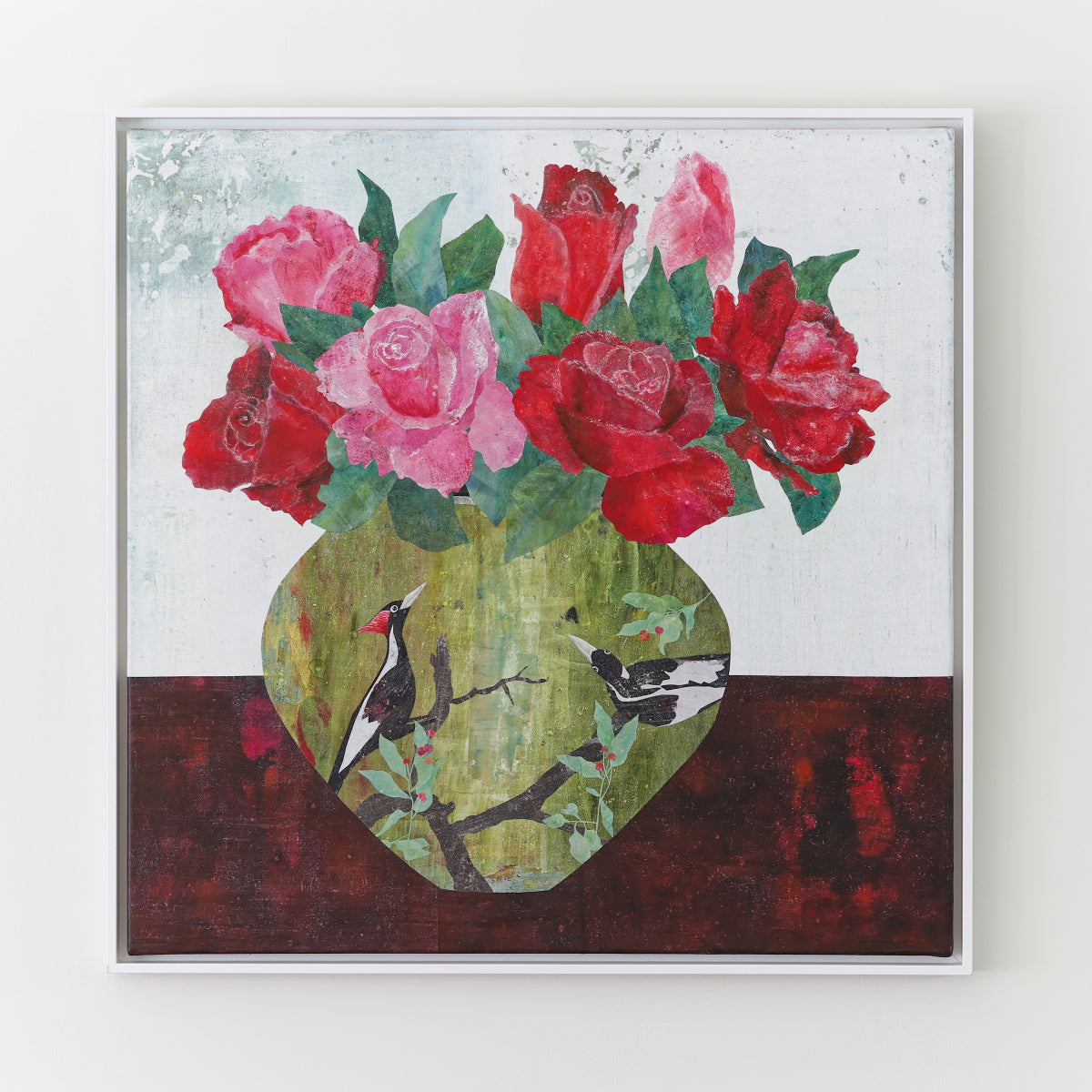 A framed square collage painting depicting a still life of red and pink roses in a green vase, with two black and white birds perched on branches, shown hanging on a light wall.