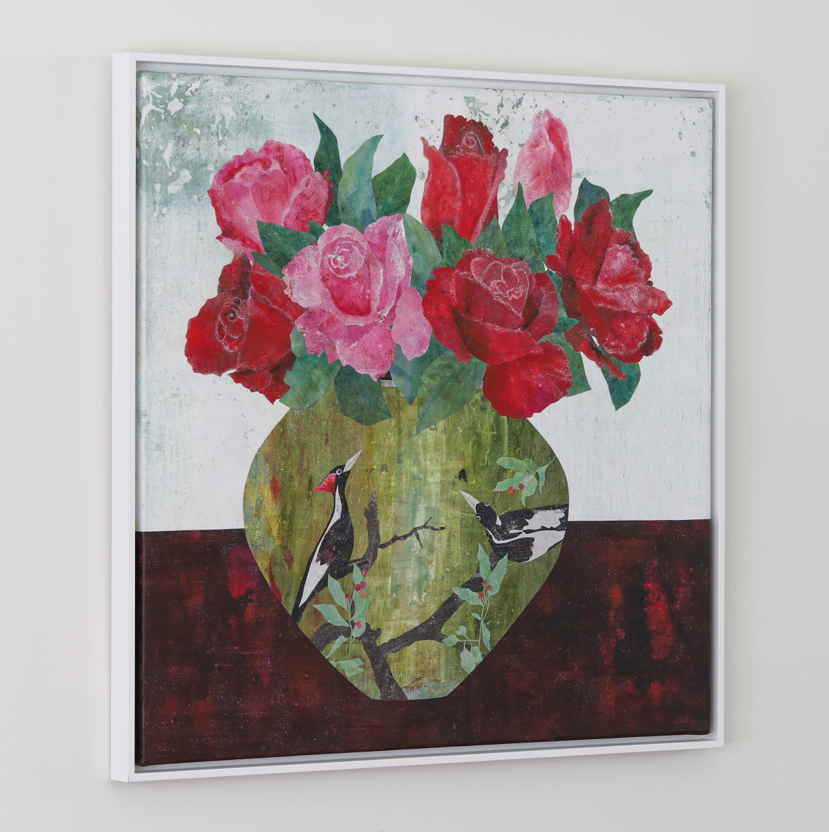 A framed square collage painting on a light gray wall, depicting a bouquet of red and pink roses in a green vase with two birds on a dark red surface against a pale background.