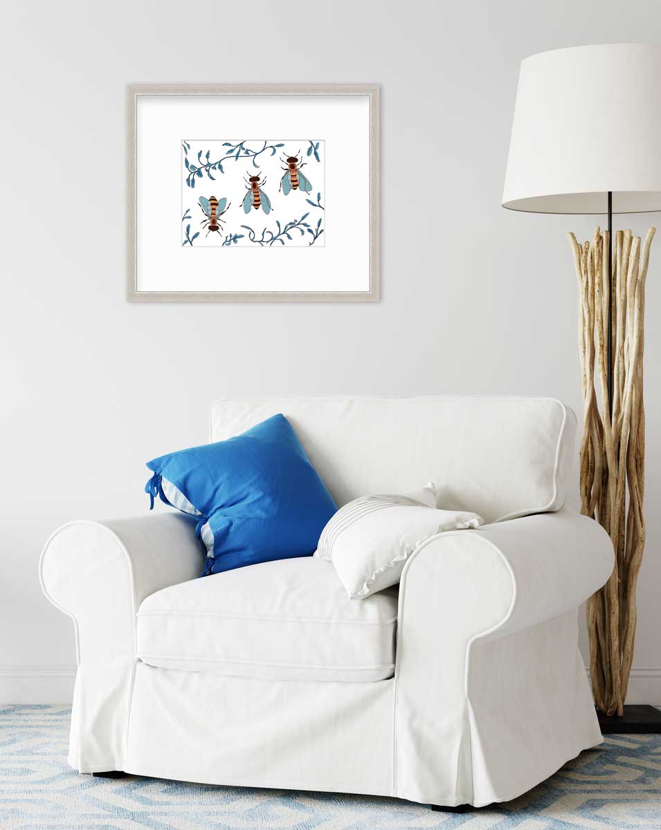 A print of three bees surrounded by blue ferns in a silver frame with a mat hangs on a white wall above a white arm chair with blue and white pillows. Behind the chair sits a floor lamp with wood sticks as the stand. 