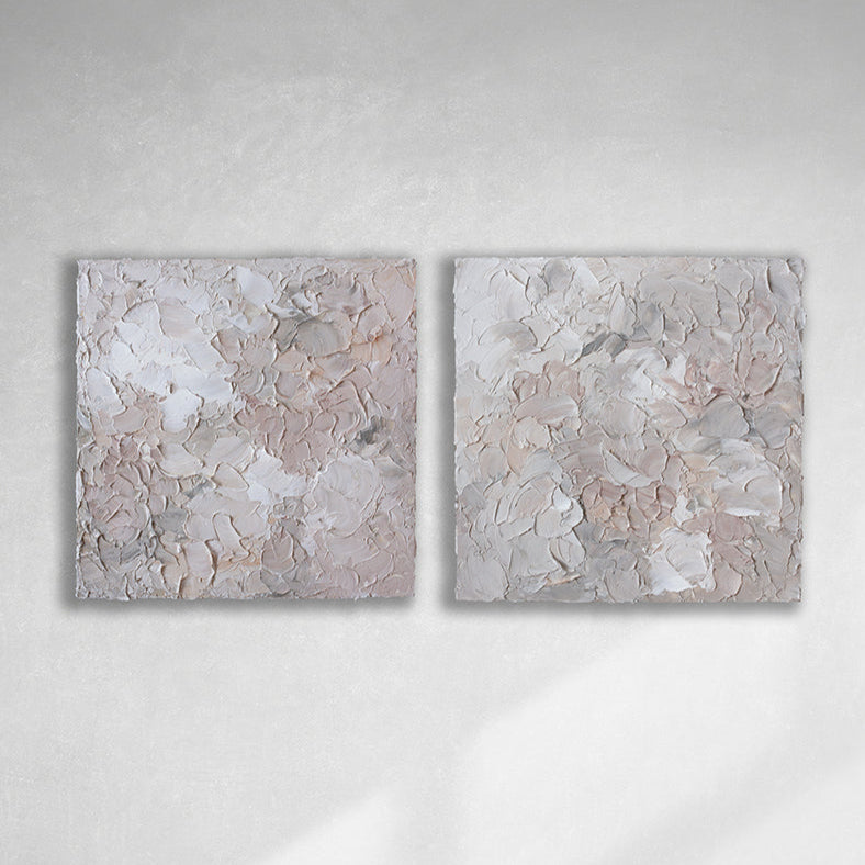 A pair of thickly textured abstract paintings in light pink, grey, coral and white by Teodora Guererra hanging on a white wall.
