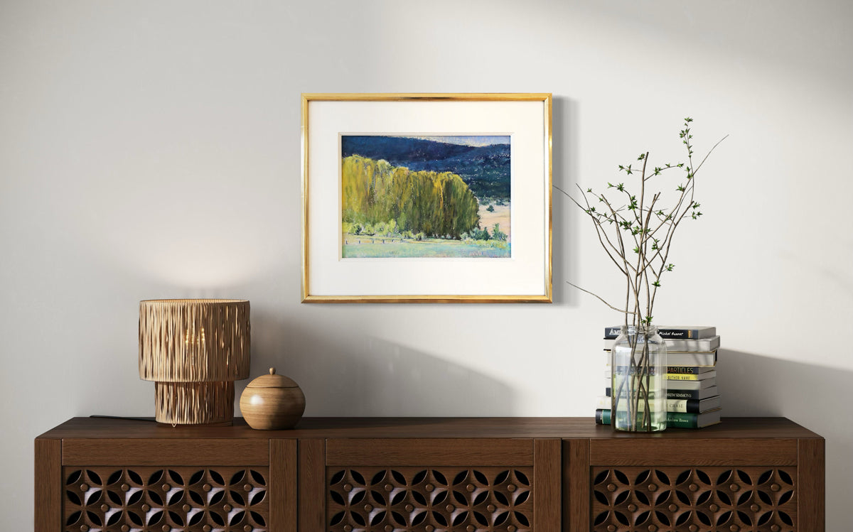 The painting, Study, Highland Greens by Ken Elliott matted in a gold frame hanging on a white wall above a dark wood credenza with decorative items and books.