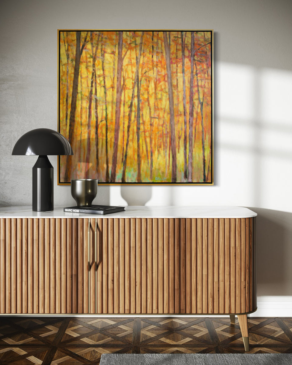An impressionistic painting similar to Wolf Kahn with tall brown trees with orange and red leaves and a warm yellow background. The painting hangs in a room over a credenza.